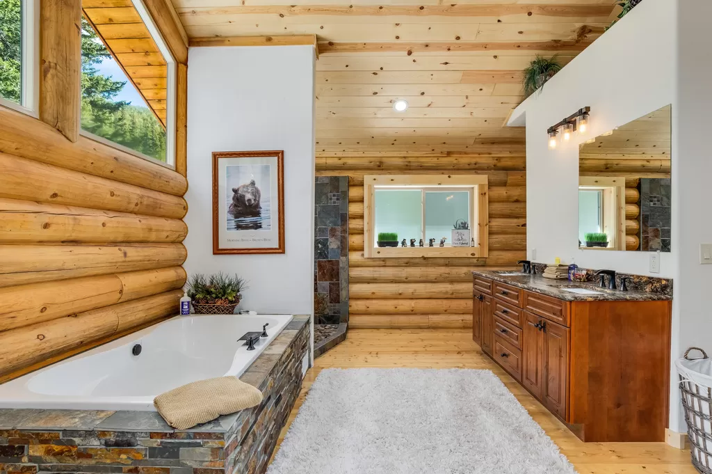 A modern log home bathroom with a log cabin aesthetic, featuring a large bathtub, stone accents, wooden walls and ceiling, dual sinks with a mirror above, and natural light from windows.