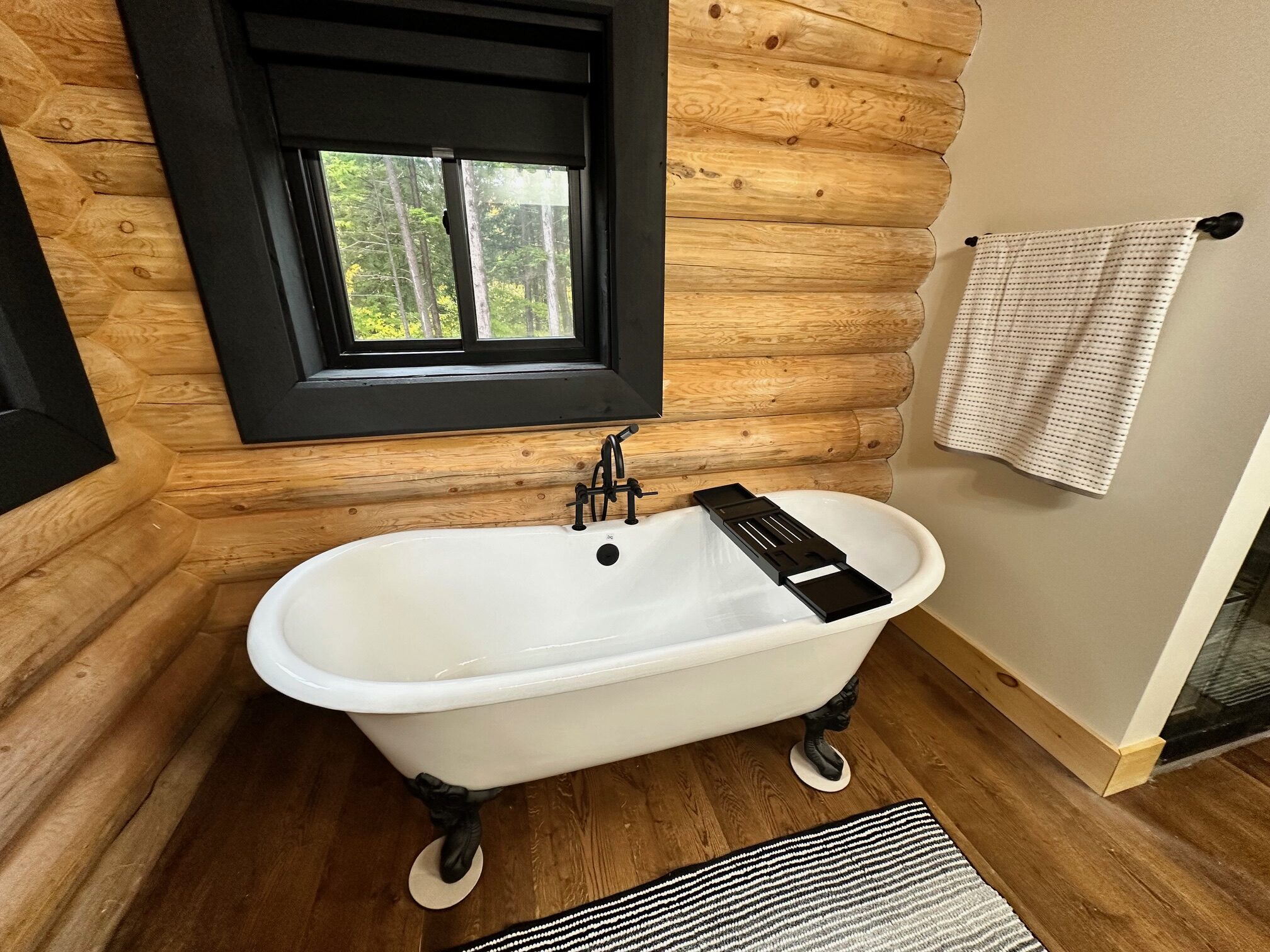 A log home white clawfoot bathtub is positioned against a wooden log wall, with a black window above it. A black and white striped bath mat lies on the wooden floor, and a towel hangs on the wall nearby.