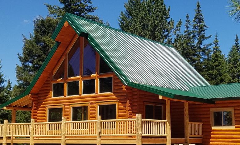 A log cabin with a green metal roof, large windows, and a wooden deck surrounded by trees.