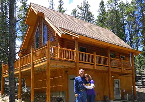 A couple stands in front of a large wooden cabin situated among trees. The cabin features multiple levels, a porch with railings, and a sloped roof.