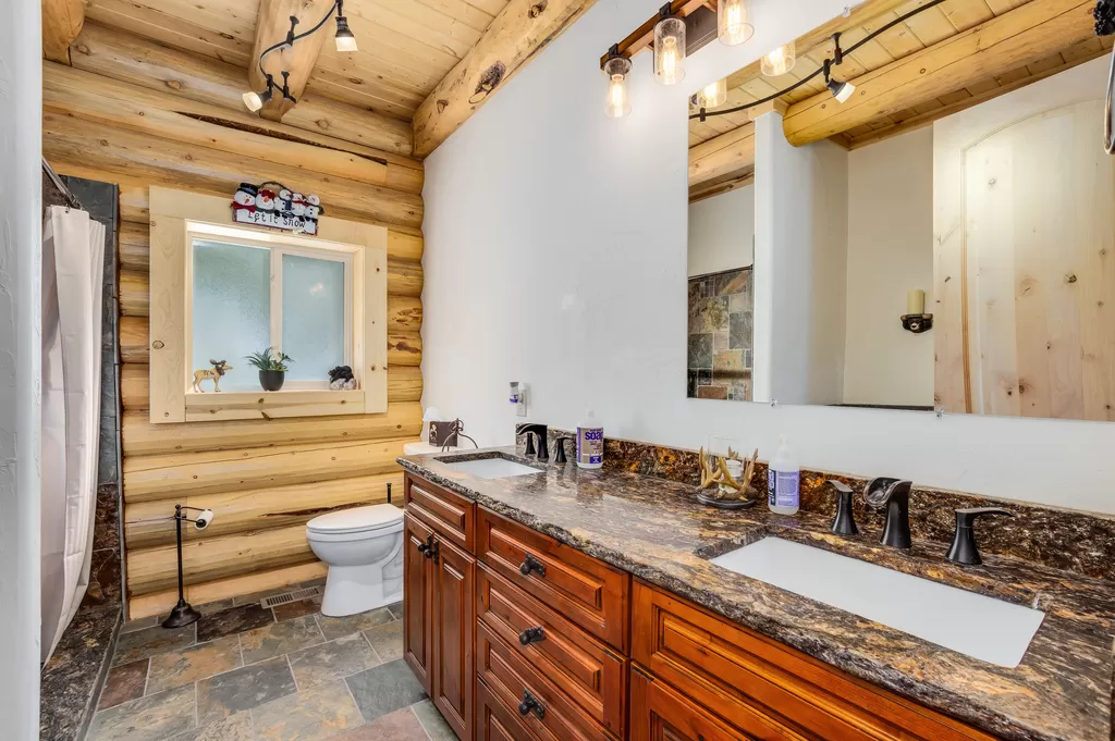 A log home bathroom with a wooden log wall, featuring a double sink vanity with dark wood cabinetry, a large mirror, and slate flooring. A shower area is visible on one side and a white toilet on the other. Natural light comes from a window above the sink area.