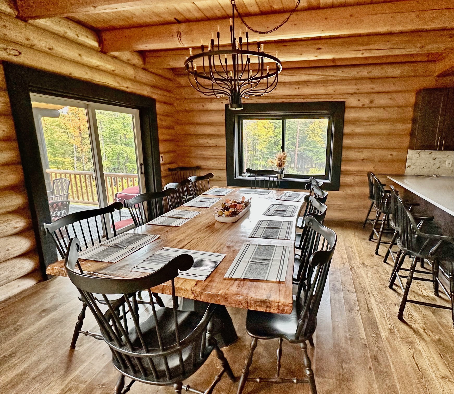 A rustic dining room featuring a large wooden table surrounded by black chairs, with a chandelier above and large windows providing a view of the outdoors. The walls are made of logs, and the floor is wooden.