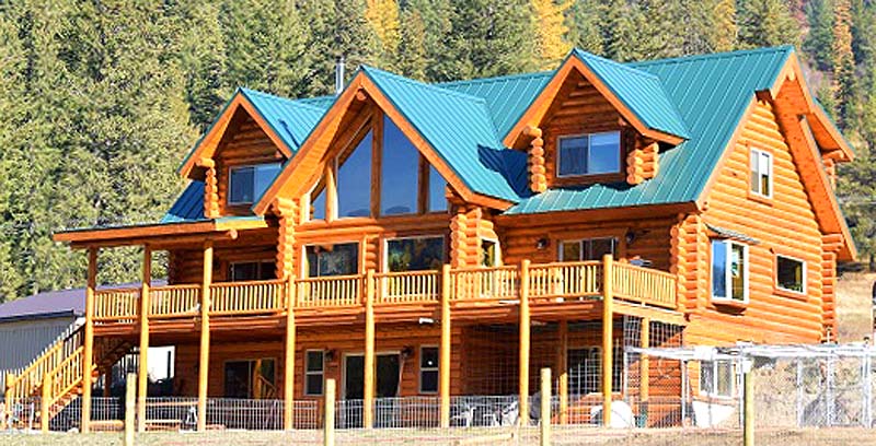 A large log cabin with a green metal roof, featuring multiple gabled sections, large windows, and a spacious wooden deck. It is surrounded by trees and natural scenery.