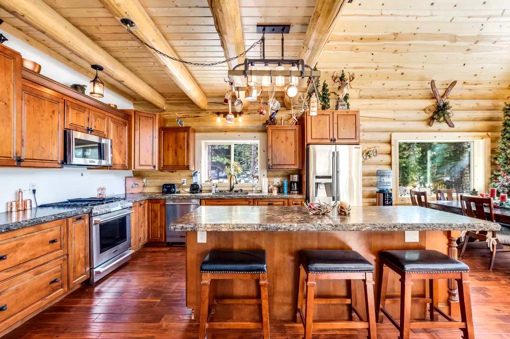 A rustic log home kitchen with wooden cabinets, a large island with bar stools, stainless steel appliances, and a wooden ceiling. Natural light enters through large windows.
