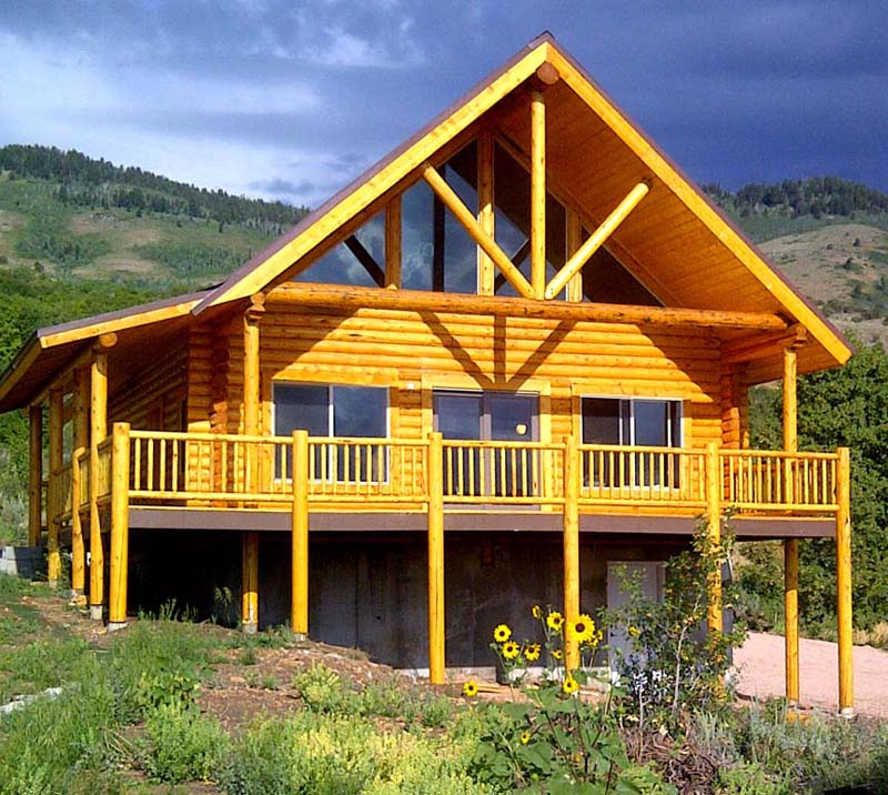 A large wooden cabin with a gabled roof, tall pillars, and a spacious deck surrounded by greenery and flowers.