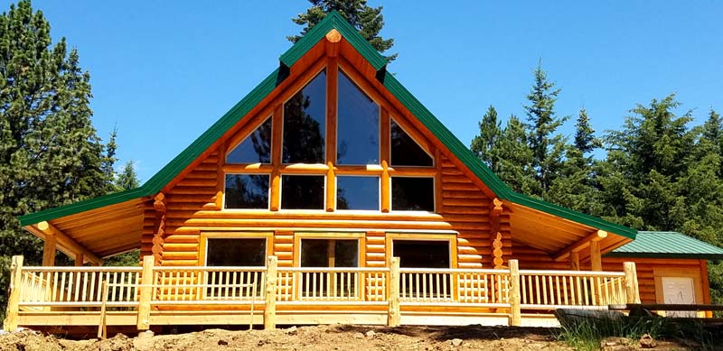 A large log cabin-style house with a green roof and multiple windows, surrounded by trees and a clear blue sky.