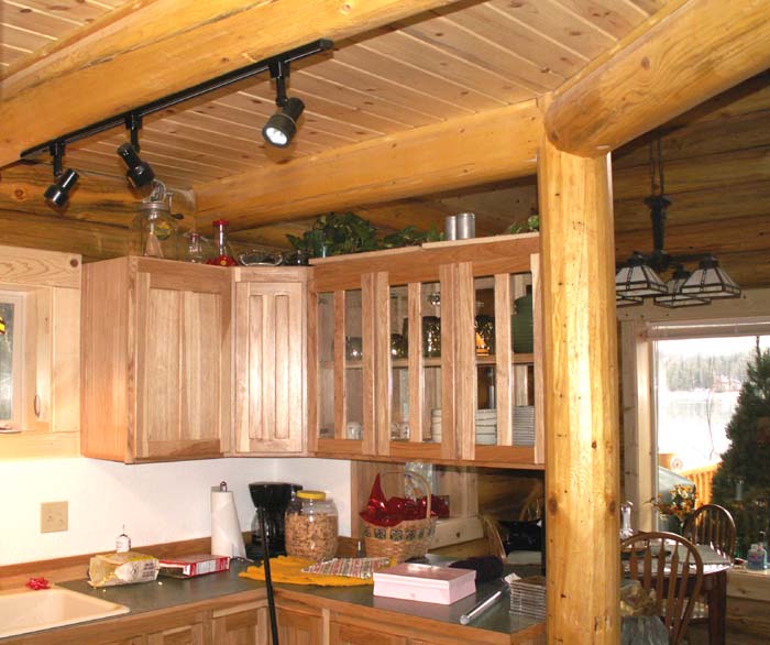 A rustic log home kitchen with wooden cabinets, a log ceiling, and track lighting. There are various kitchen items visible on the counter and shelves.
