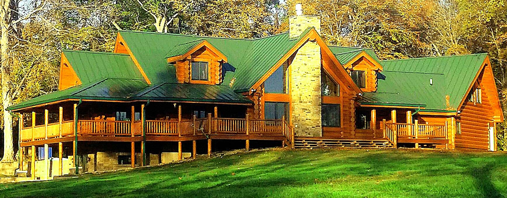 A large wooden cabin with green roofing, featuring multiple gables, a stone chimney, and a wraparound porch situated in a lush green landscape.