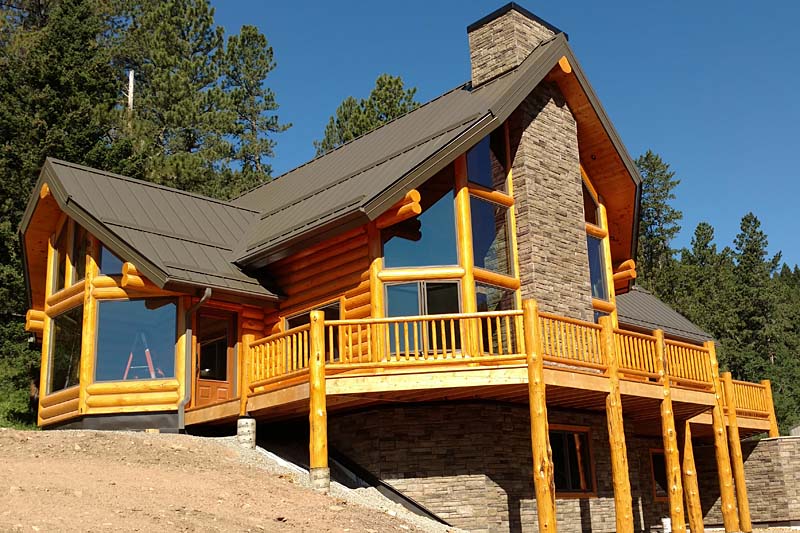 A large log cabin with a stone chimney, featuring expansive glass windows and a wooden deck surrounded by trees and blue sky.