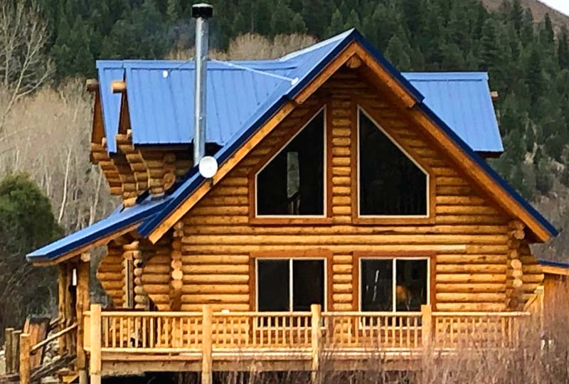 A wooden log cabin with a blue metal roof, featuring large windows and a deck. It is surrounded by greenery and mountains in the background.