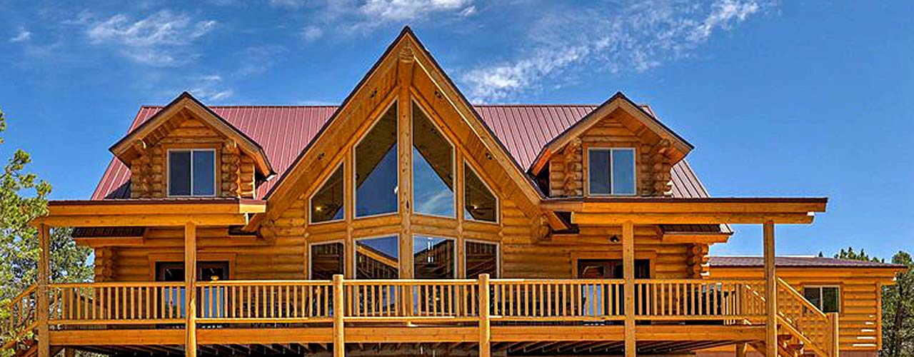 A log cabin with a steeply pitched roof, large windows, and a wooden deck under a clear blue sky.