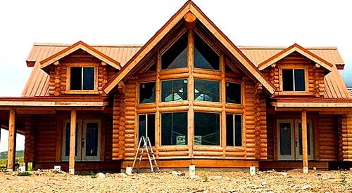 A log cabin-style house with a large front porch, featuring a triangular roof and extensive windows. A ladder is positioned in front of the house.