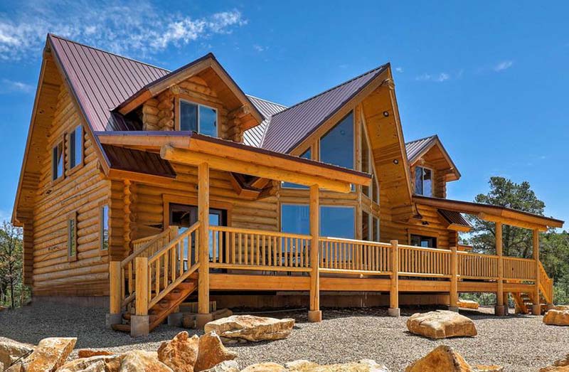 A large log cabin-style house with a wooden deck and expansive windows, set against a blue sky.
