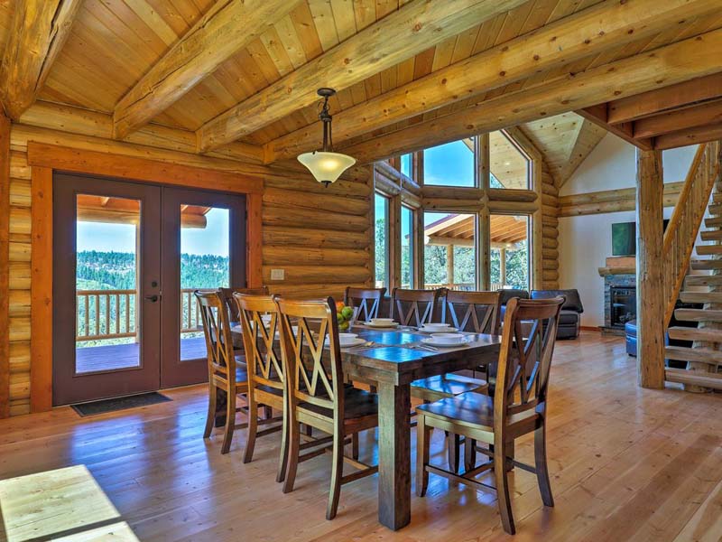 A rustic dining area with a large wooden table and several chairs, surrounded by log walls and large windows that offer a view of the outdoors. There is a staircase leading to an upper level in the background.