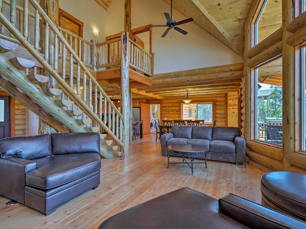 A spacious interior of a log home featuring wooden beams, a staircase, dark leather furniture, large windows, and an open layout connecting to a dining area.