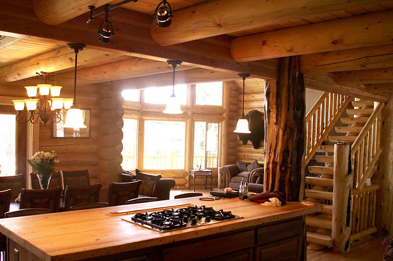 A rustic log home kitchen with a wooden island, gas stove, and exposed log supports. Large windows let in natural light, revealing a cozy living area with a couch and staircase in the background. A chandelier hangs over the dining table. Cascade island kitchen into great room view 800