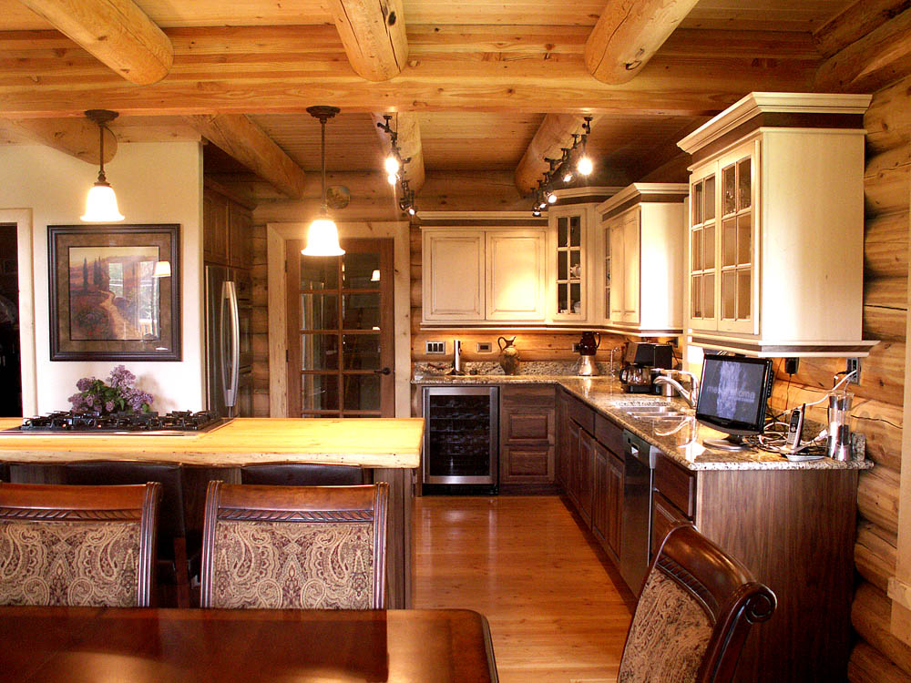 A rustic log home kitchen with wooden beams and cabinets, featuring a granite countertop and modern appliances, adjacent to a dining area.