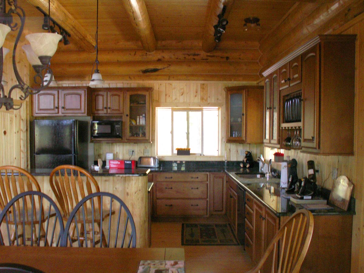 A wooden log home kitchen with a rustic design, featuring dark cabinetry, a window letting in natural light, and a dining table with wooden chairs.