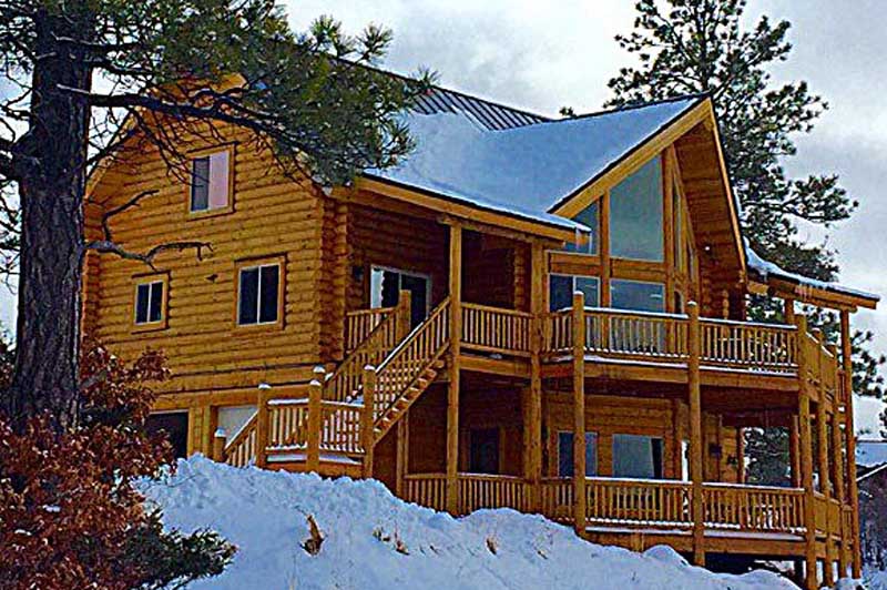 A large wooden log cabin with multiple levels, surrounded by snow and trees. The cabin features a sloped roof and expansive windows, giving it a modern yet rustic appearance.