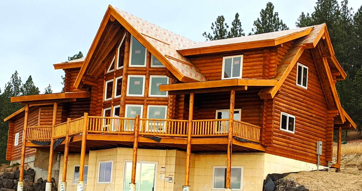 A large, multi-story log cabin with a peaked roof, featuring numerous windows and a spacious balcony on the front. The cabin is set against a backdrop of trees.