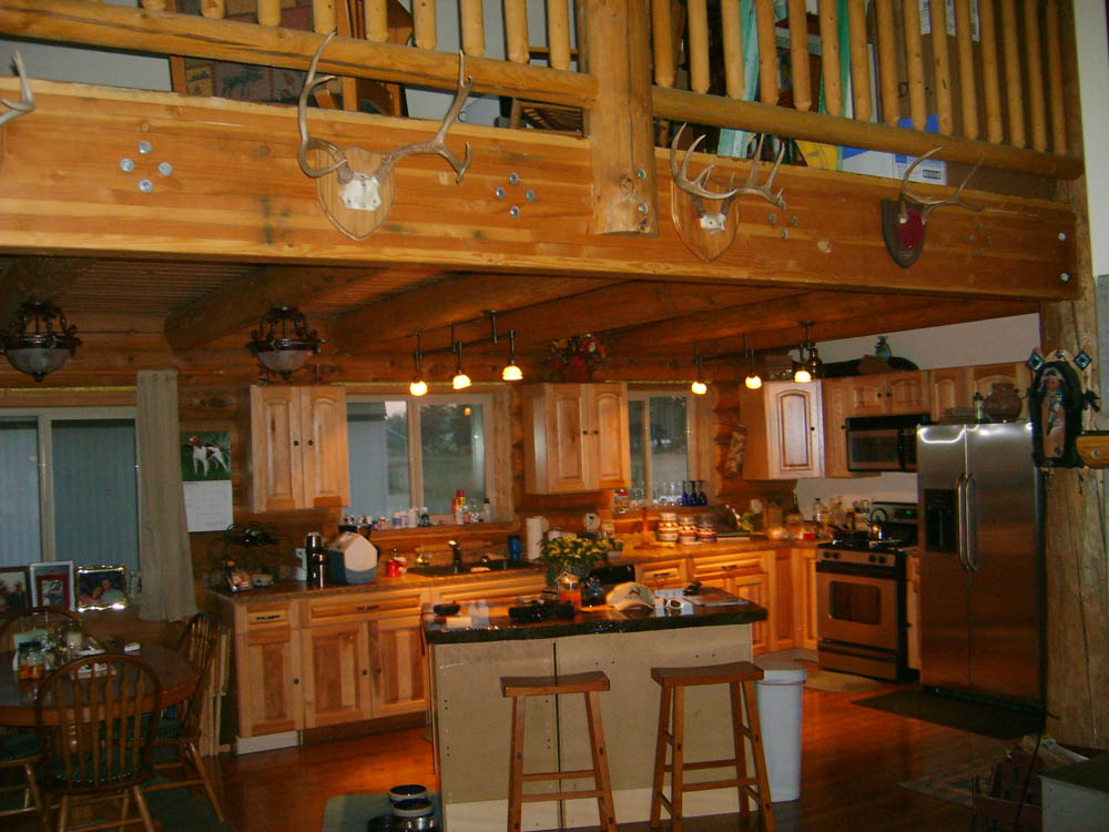 A wooden log home kitchen with a spacious layout featuring a central island, wooden cabinetry, and appliances. There are beams in the ceiling and some decorative items on display.