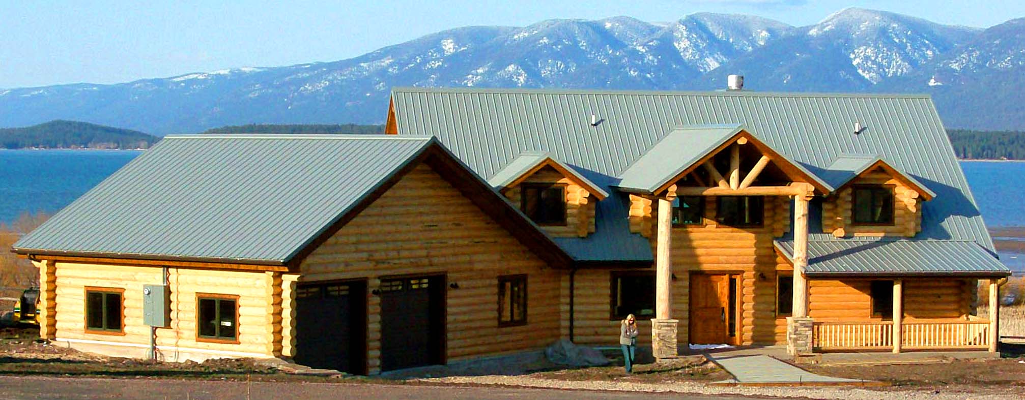 A large log cabin-style house with a metal roof, situated near a body of water and mountains in the background.