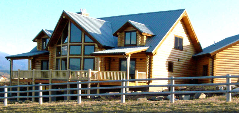 A large log cabin with a modern design, featuring a mix of wood and glass elements, set against a clear blue sky. The house has a prominent porch area and is surrounded by an open field and fence.