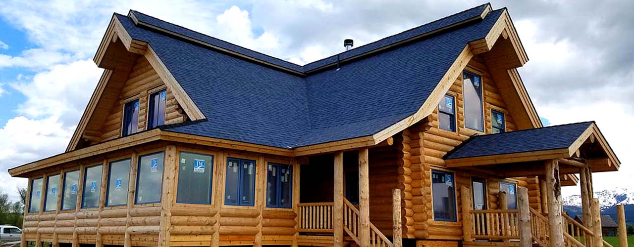 A large log cabin with a blue roof and multiple windows, featuring a porch and surrounded by grassy areas under a partly cloudy sky.