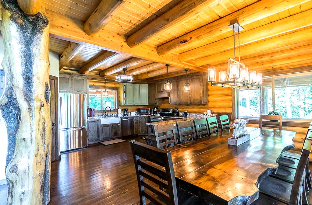 A spacious wooden cabin interior featuring a large dining table, log beams on the ceiling, and an open kitchen area with modern appliances.