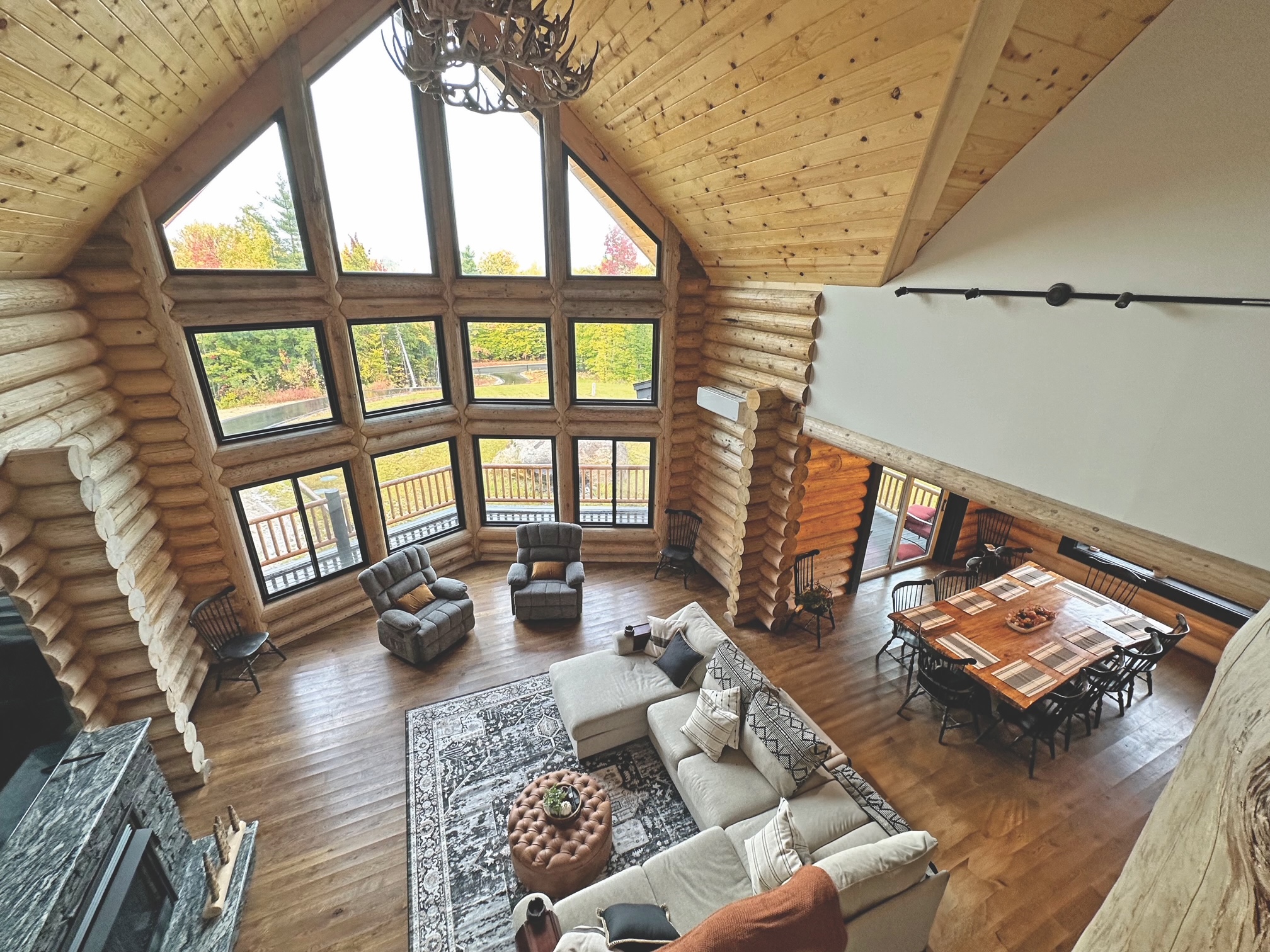 A spacious log home interior with large windows, wooden beams, and a cozy living area featuring comfortable furniture and a dining table.