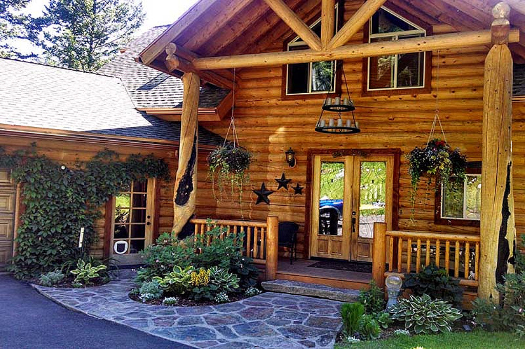 A rustic log cabin-style home with wooden pillars, a stone pathway, hanging plants, and a lush garden.