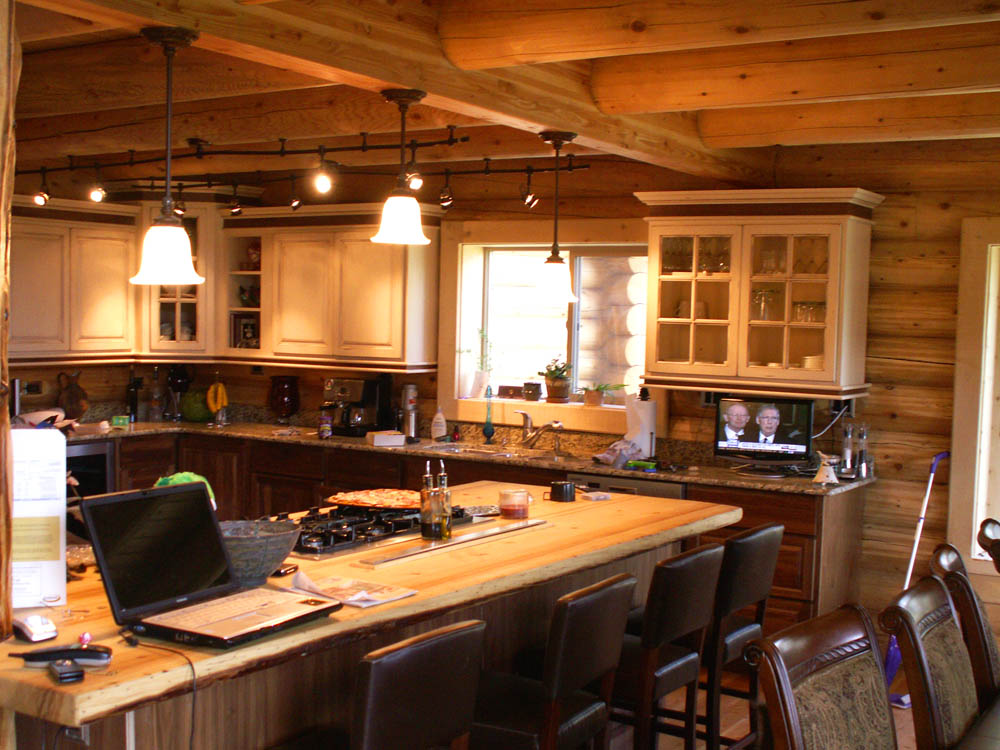 A rustic log home kitchen with wooden beams and cabinets, featuring a central island with bar stools, a stove, and various kitchen appliances. There is a window allowing natural light in, and a television displaying two people in the background.