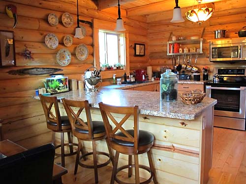 A rustic log home kitchen with wooden log walls, featuring a countertop bar with three stools, a window letting in natural light, and various kitchen appliances like an oven and microwave. Decorative plates hang on the wall, and there's a fish tank on the counter.