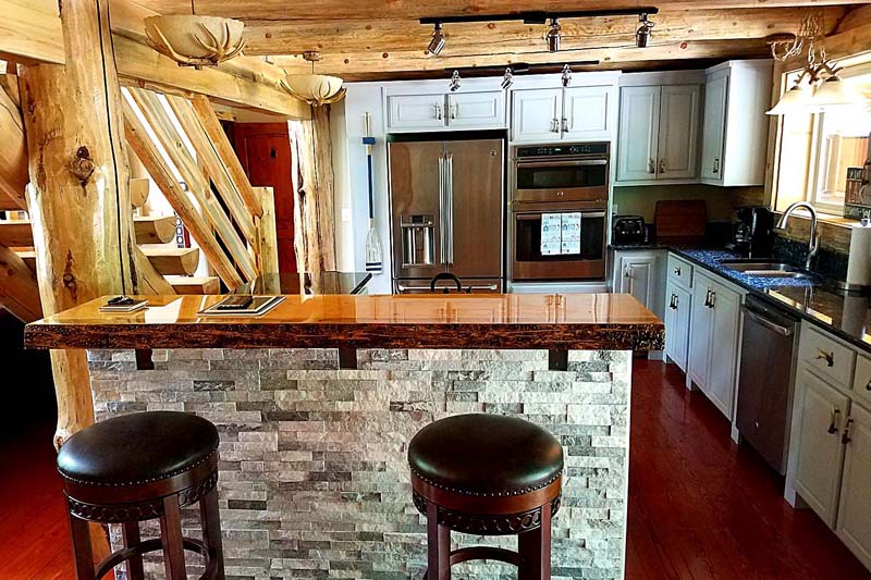 A rustic log home kitchen with wooden beams, a stone bar counter, and modern appliances. There are two stools at the bar, and the overall design combines natural elements with contemporary features.