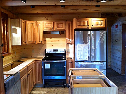 A log home kitchen with wooden cabinets, a stainless steel refrigerator, a stove, and countertops. The kitchen is partially finished with some items still wrapped in plastic.