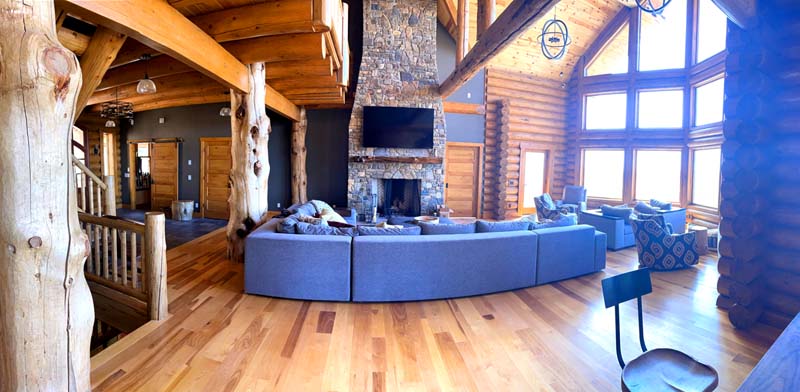 A spacious, rustic log home interior with a large stone fireplace, wooden beams, and panoramic windows. A gray sectional sofa is in the center, surrounded by wooden flooring.