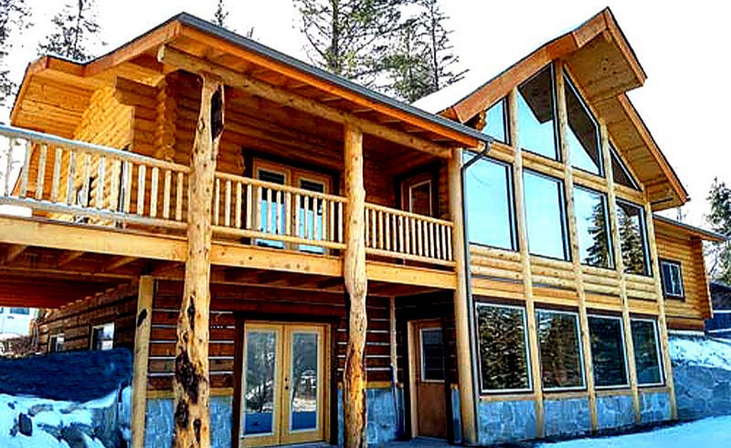 A wooden cabin with large windows, a balcony, and a sloped roof set in a snowy landscape surrounded by trees.