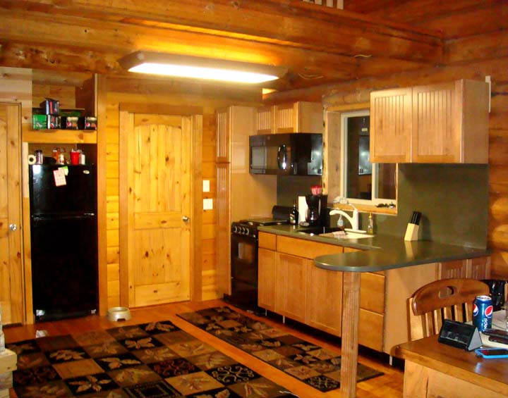 A rustic log home kitchen with wooden cabinets, a black refrigerator, and a gas stove. There is a sink and countertop space, along with various appliances. The floor features a patterned rug, and the walls are made of logs.