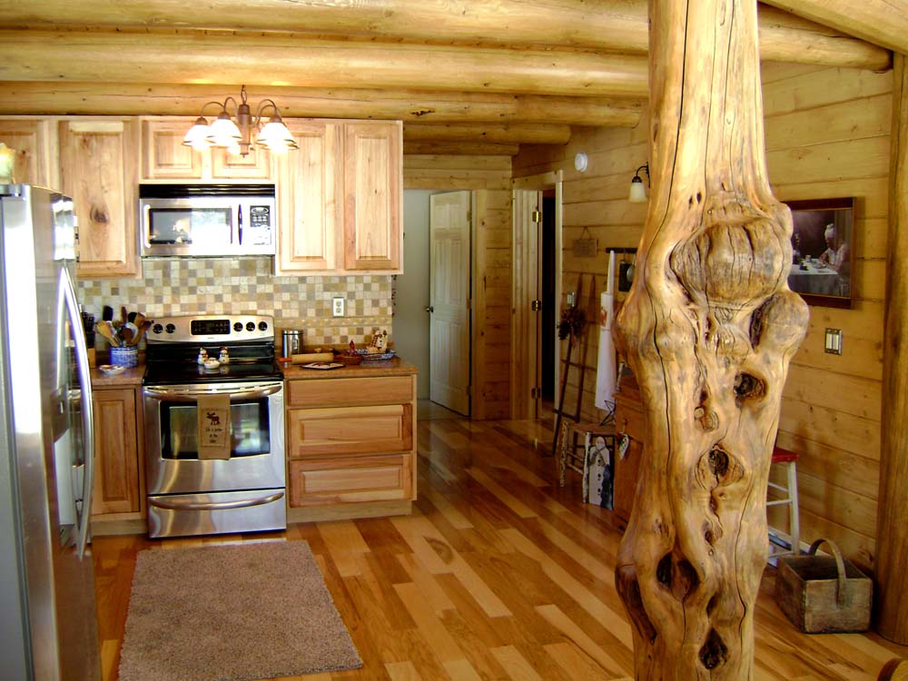 A rustic log home kitchen with wooden cabinets, stainless steel appliances, a large wooden support beam, and a cozy atmosphere.