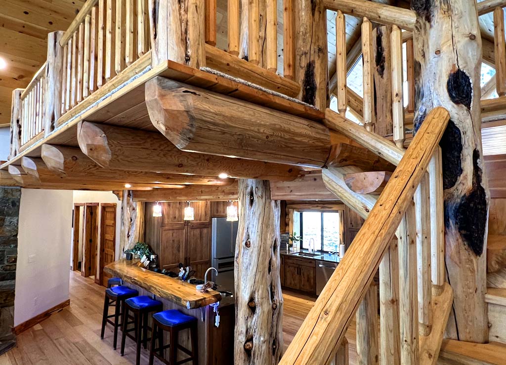 A rustic log home wooden interior of a log cabin with a balcony overlooking the living area and a kitchen visible in the background. The staircase is made of wood, and there are barstools at a counter made from large logs.