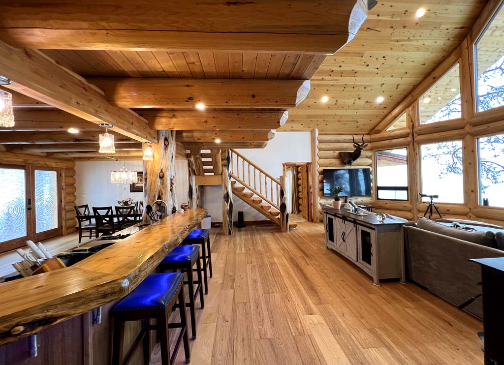 Interior of a rustic log home cabin with wooden beams, a staircase, stone fireplace, and dining area featuring wooden furniture. Large windows allow natural light to flood the space.