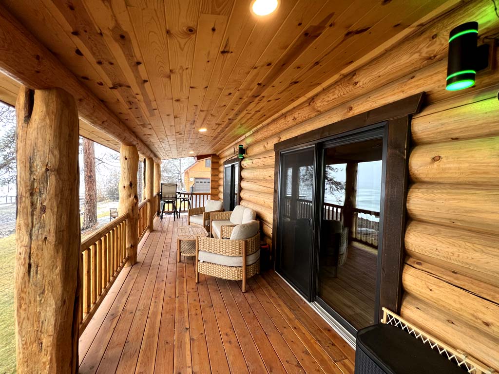 A wooden porch featuring log cabin-style walls, two chairs, and large sliding glass doors leading into the interior.