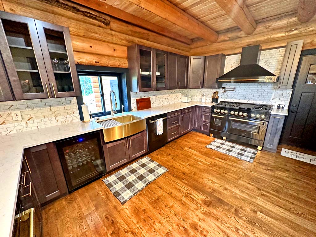 A spacious log home kitchen featuring wooden beams, dark cabinetry, a large sink with a farmhouse-style faucet, and an oven range. The countertops are made of white material with speckles, complemented by stone backsplash. Two plaid mats are placed on the floor in front of the sink and stove.