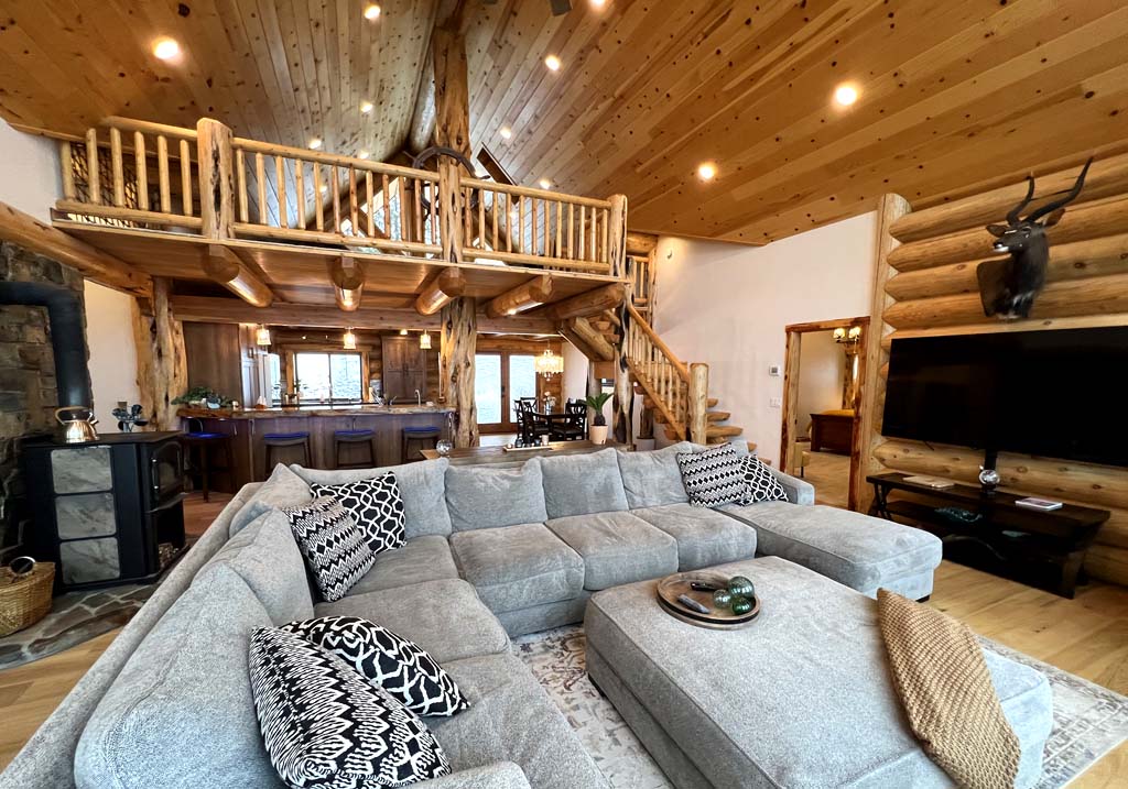 A spacious log home interior featuring a large gray sectional sofa with decorative pillows, wooden ceiling beams, and a staircase leading to an upper level. The space is well-lit with natural light and includes a stone wall and wooden accents throughout.