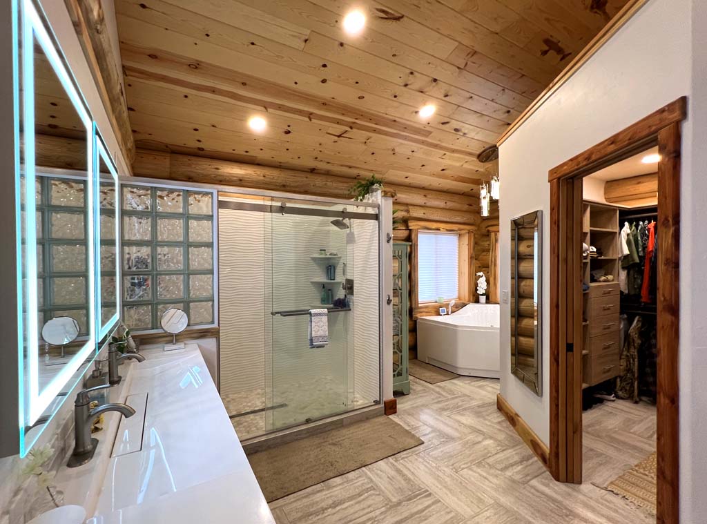 A modern log home bathroom with wooden log accents, featuring a glass shower enclosure, a freestanding bathtub, and double sinks with an illuminated mirror.