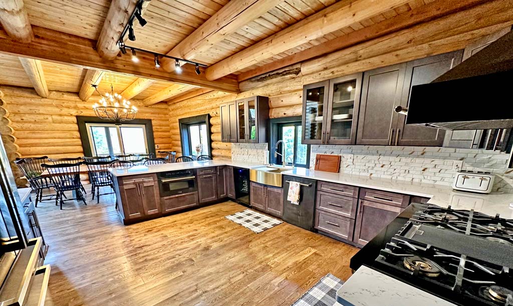 A spacious log home kitchen with wooden beams and cabinetry, featuring a large window, a farmhouse sink, and modern appliances. A dining area is visible in the background with a chandelier and several chairs.