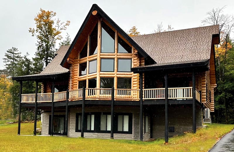 A large log cabin with large windows and a sloped roof, situated on a grassy area surrounded by trees.