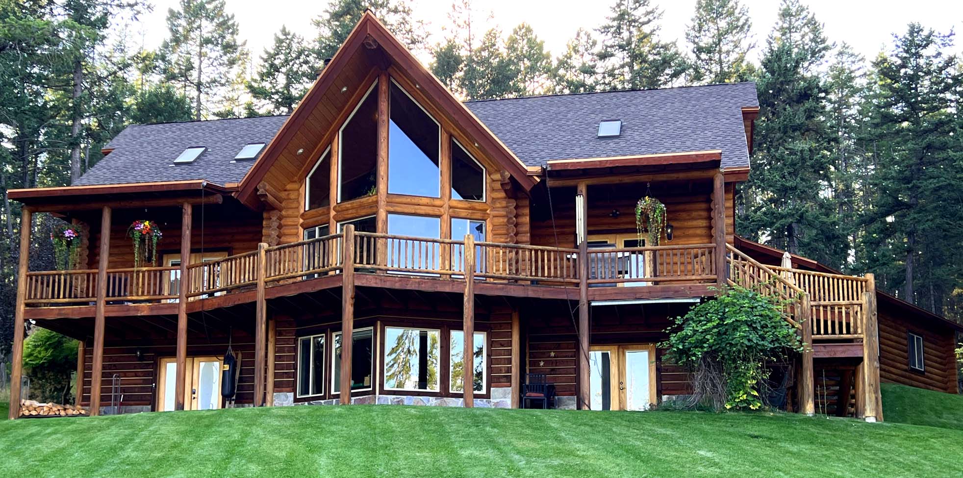 A large log cabin-style house with multiple levels, large windows, and a wraparound porch set in a grassy area surrounded by trees.