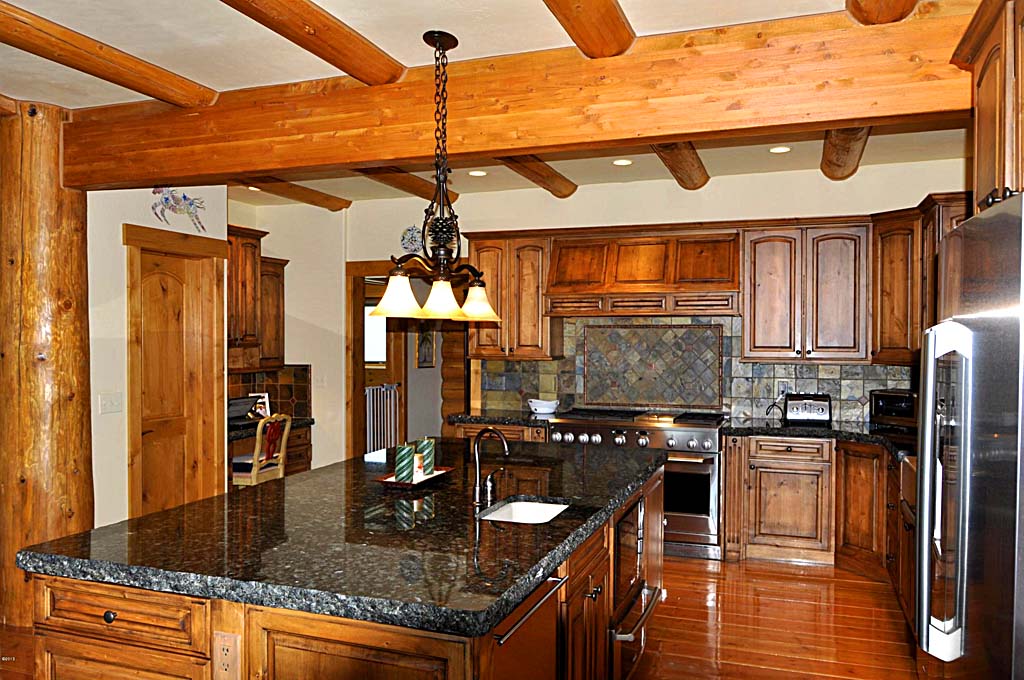 A modern log home kitchen featuring wooden beams, dark cabinetry, and a large granite island with a built-in sink.