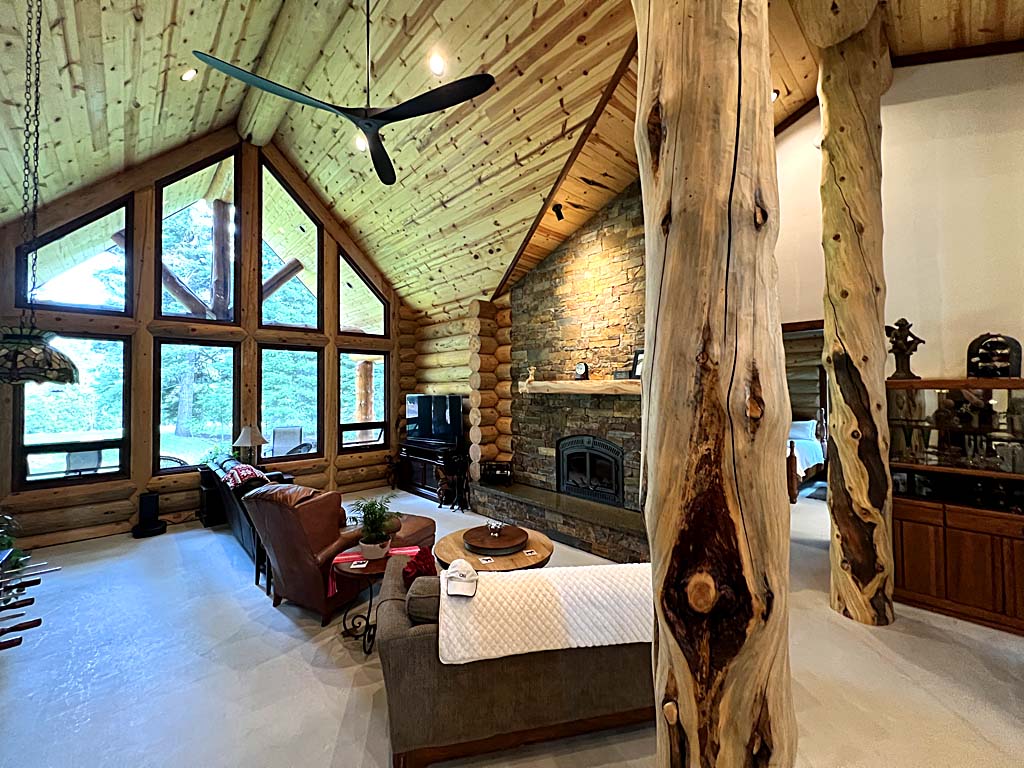 A cozy log home interior of a log cabin featuring large windows, wooden beams, and a stone fireplace. The room contains comfortable seating arrangements and decorative elements like a coffee table and wall décor.